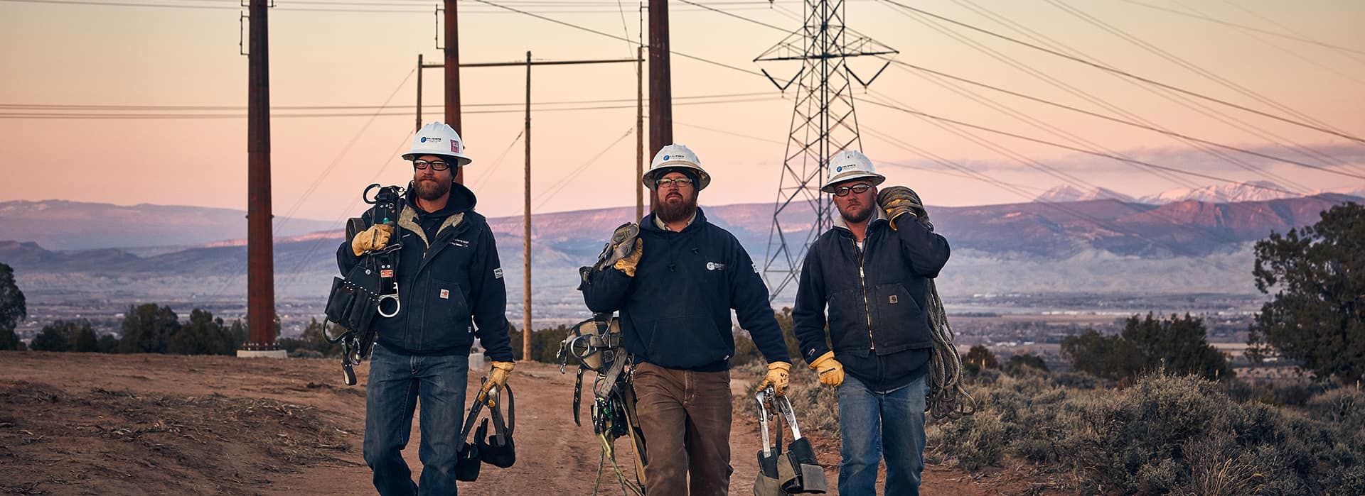 Three linemen carrying their gear along a ridge at dusk, transmission towers behind them